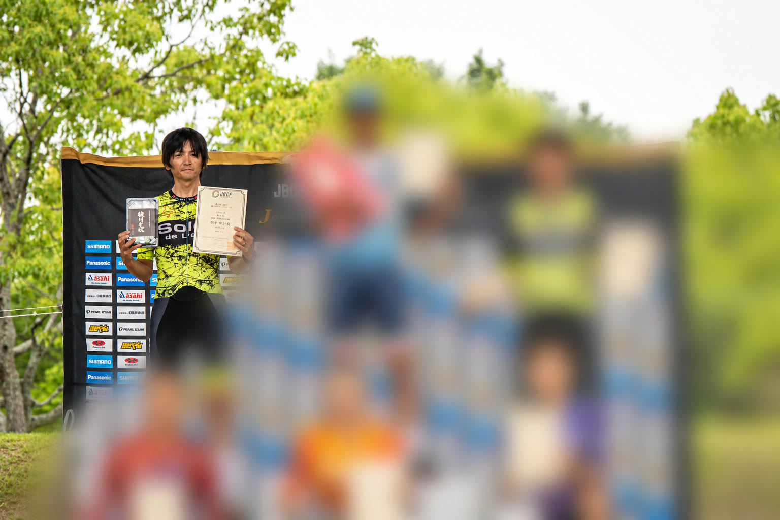 表彰台の左上で賞状を手にするのりりん。ほかの参加者はガラス風にぼかされている